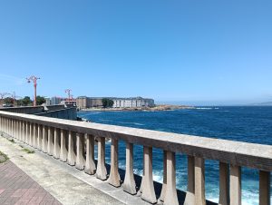 A couple of buildings over the cost with the ocean viewed from a street that features a stone railing on a clear day.