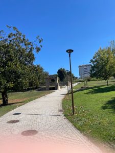 Brick-paved pathway through a green park with trees, street lamps, a stone archway, and a modern building under a clear blue sky.