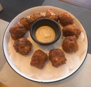 plate of ten golden-brown fried chicken bites arranged in a circle around a small black bowl of creamy dipping sauce, served on wax paper liners.
