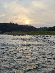 The setting sun glows above the tree-covered landscape at Kariyathumpara, Kozhikode, Kerala, while a flowing river fills the foreground. The warm reflection on the water creates a peaceful evening scene.
