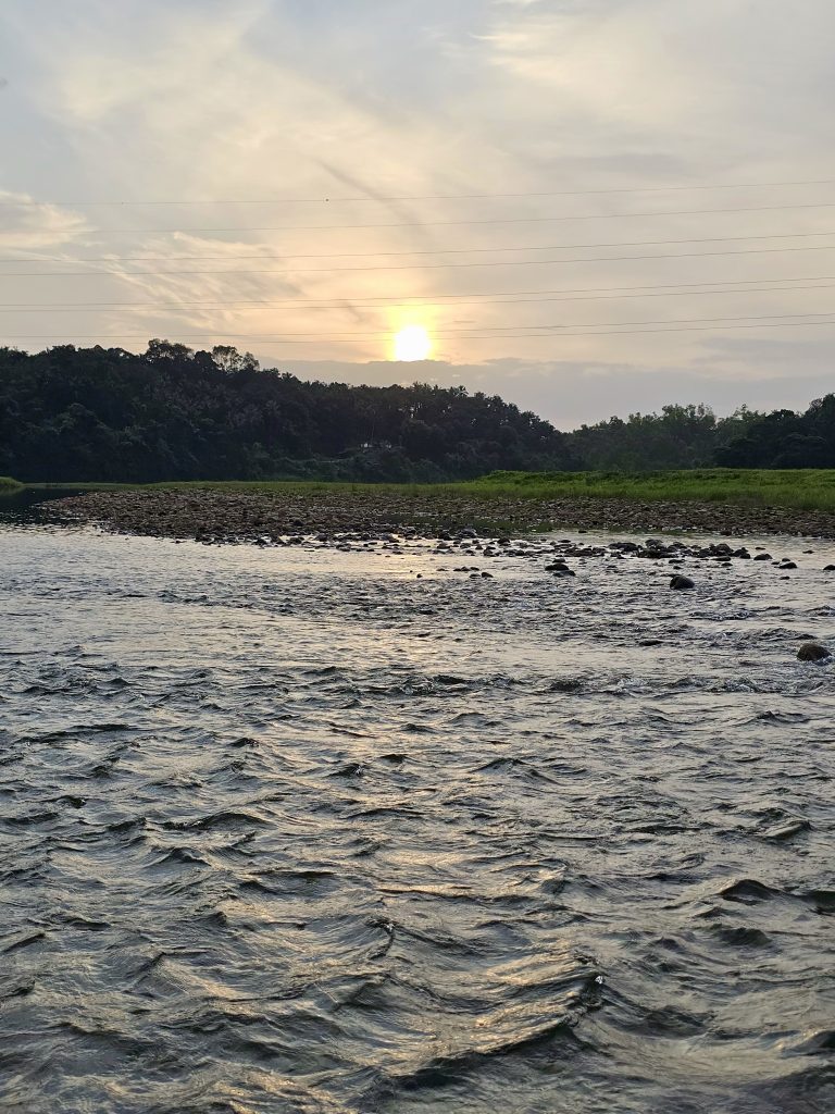 The setting sun glows above the tree-covered landscape at Kariyathumpara, Kozhikode, Kerala, while a flowing river fills the foreground. The warm reflection on the water creates a peaceful evening scene.