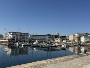 Panoramic view of A Coruña marina with boats reflected in calm water under a clear blue sky.