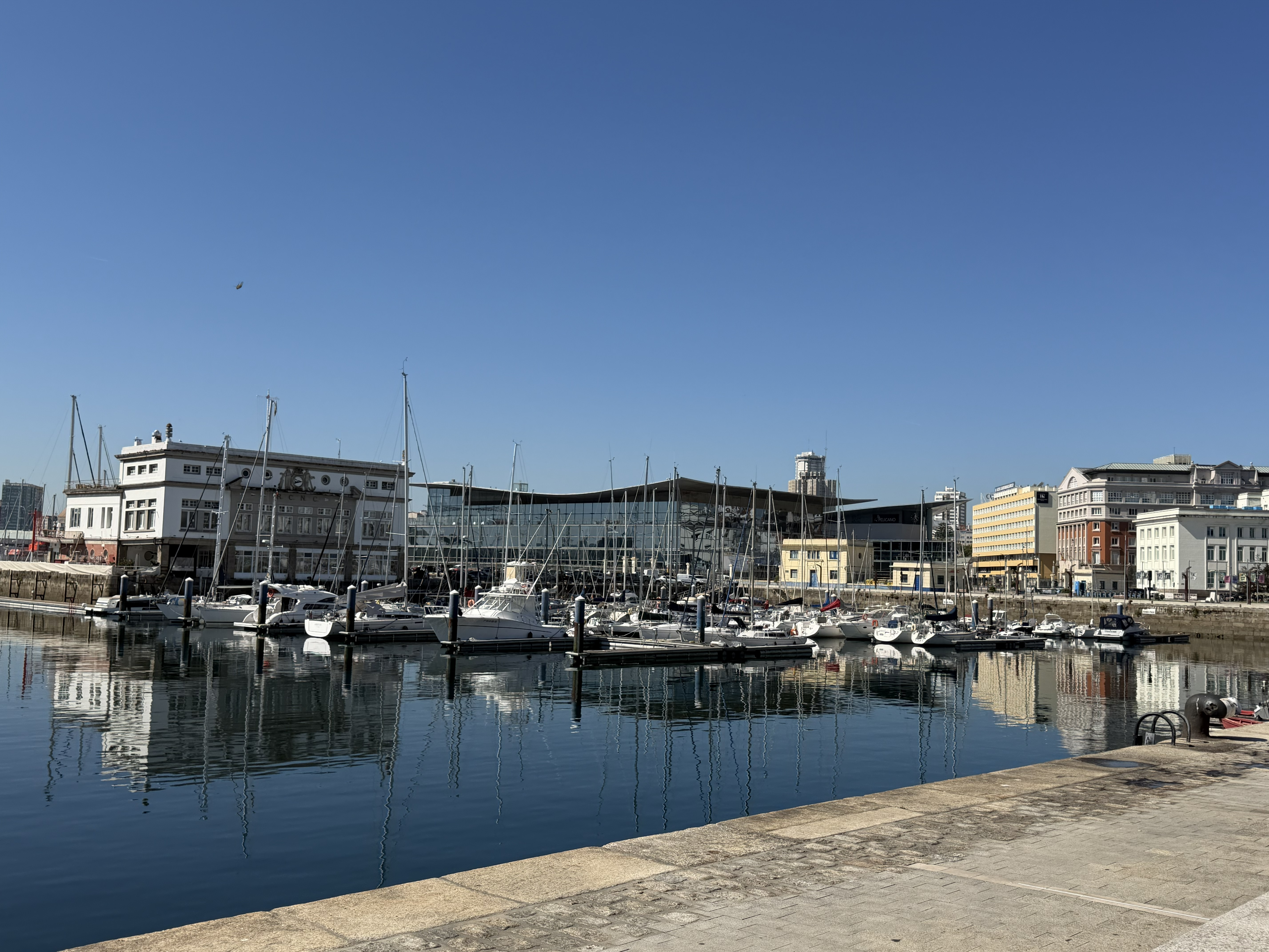 Panoramic view of A Coruña marina with boats reflected in calm water under a clear blue sky.