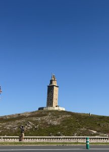 Hercules Tower on a green rocky hill under a clear blue sky, with a road and a person viewing the lighthouse.