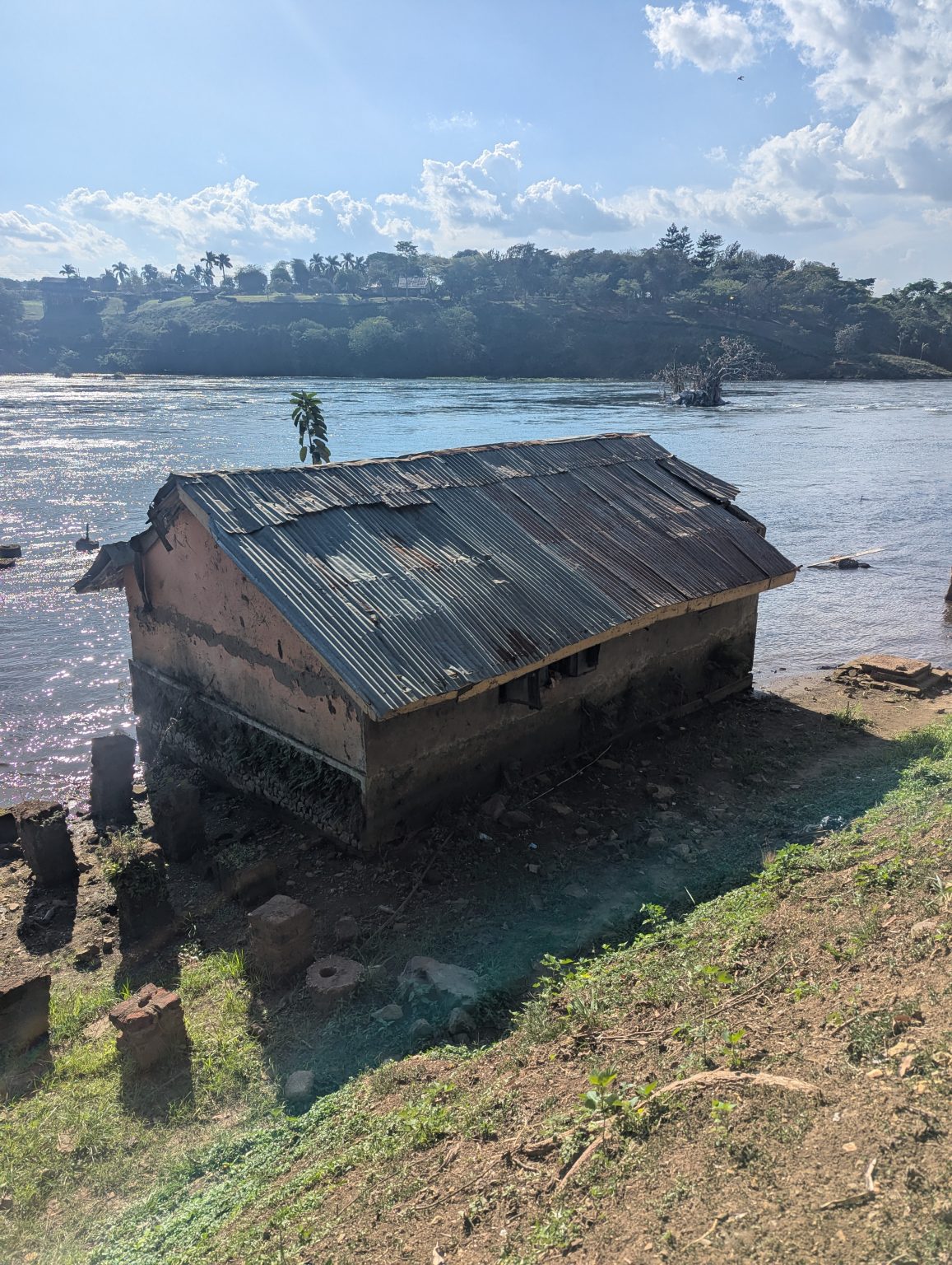 Weathered building with a rusty corrugated roof beside a river, surrounded by grass, rocks, and distant green hills.