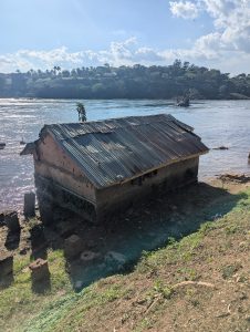 Weathered building with a rusty corrugated roof beside a river, surrounded by grass, rocks, and distant green hills.
