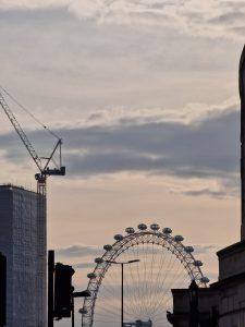 Moody evening silhouette of a tall construction crane and the London Eye ferris wheel set against a warm sunset sky.