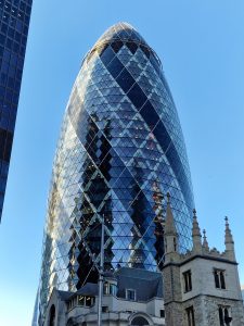 Modern glass curved London skyscraper rising behind a traditional stone church tower under a clear blue sky.