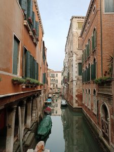 Narrow Venetian canal lined with historic orange and brick buildings, green window shutters, and small moored boats reflecting in calm water.