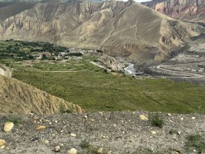 A wide landscape view looking down into a valley where green patches of farmland and a rural village rest beside a winding river. Massive, heavily textured tan cliffs and mountains rise sharply around the valley floor.