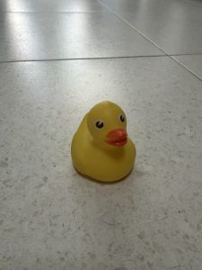 Yellow rubber duck with a bright orange beak and large black eyes on a light speckled tiled floor, viewed from above.