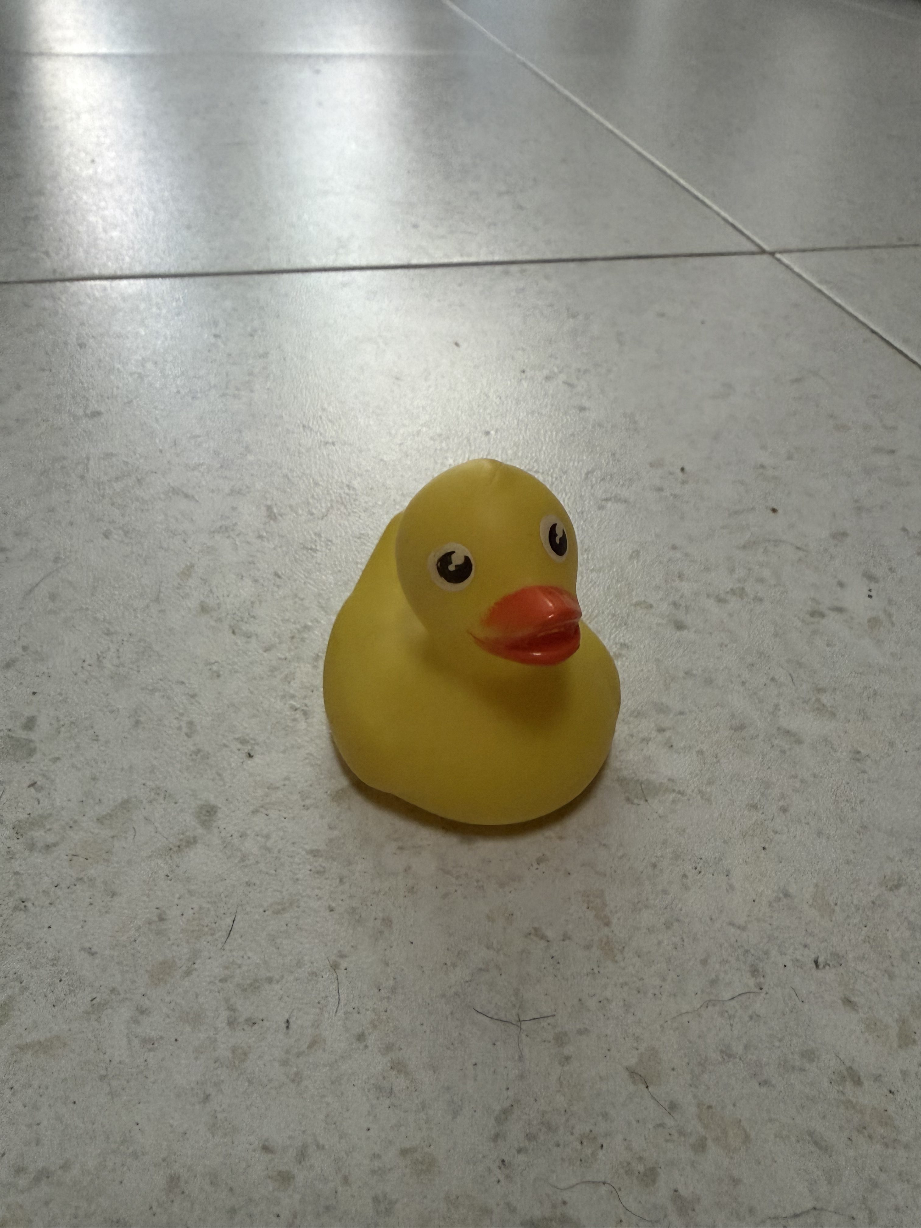Yellow rubber duck with a bright orange beak and large black eyes on a light speckled tiled floor, viewed from above.
