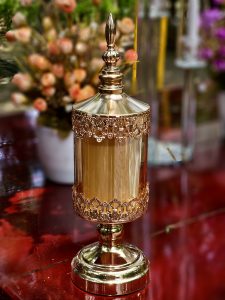 A decorative glass candle holder with a golden finish stands prominently against a softly blurred garden background, creating an elegant still-life composition. Location: Ayamkudy, Kottayam, Kerala.