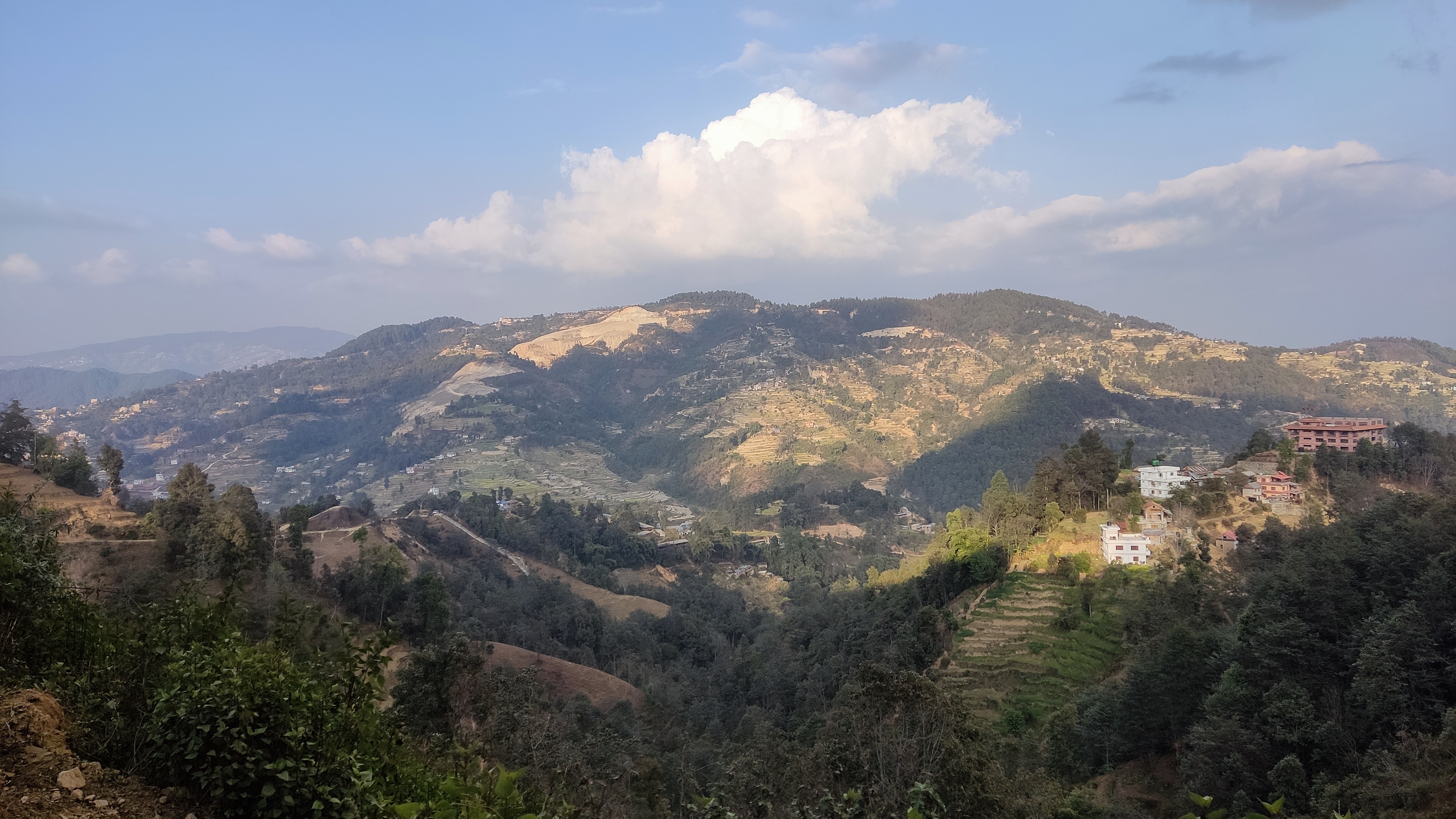 A panoramic view of a mountainous landscape under a partly cloudy sky. Rolling hills are covered with patches of lush greenery and terraced fields, showing various shades of brown and green