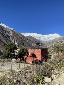 A traditional red and gold Tibetan Buddhist monastery (gompa) set against a stark mountain valley with a sweeping wall of snow-capped mountains in the background.