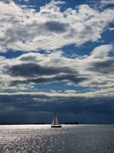 Silhouette of a small sailboat sailing on glistening ocean water beneath dramatic white clouds.