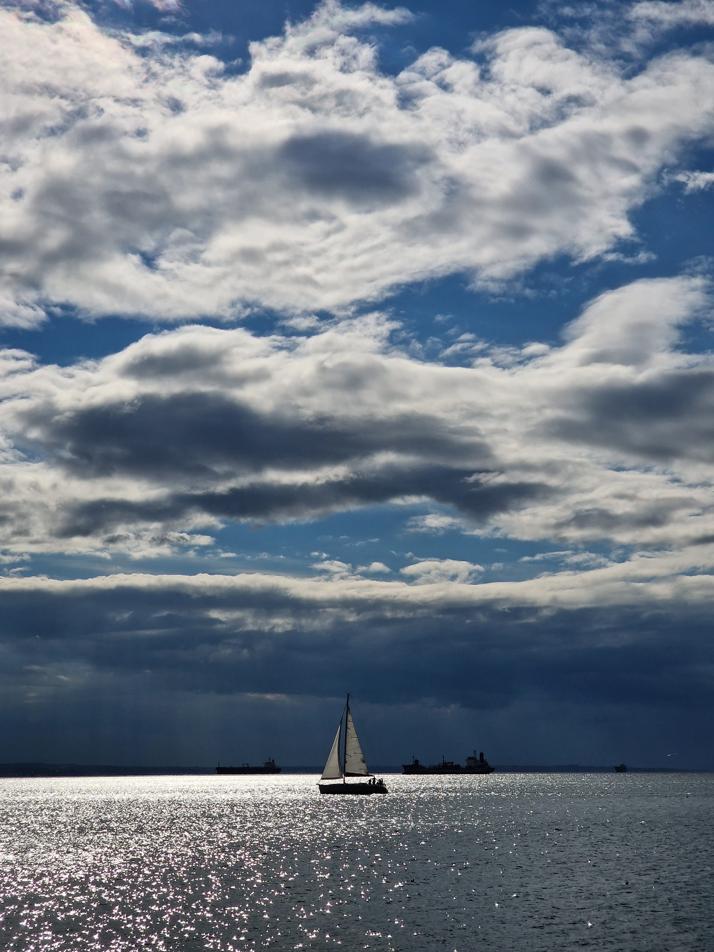 Silhouette of a small sailboat sailing on glistening ocean water beneath dramatic white clouds.