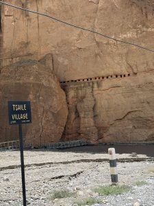 A sign reading "TSAILE VILLAGE 1 K.M." standing beside a rocky riverbed with a cliff face in the background featuring a line of ancient, hand-carved sky caves.