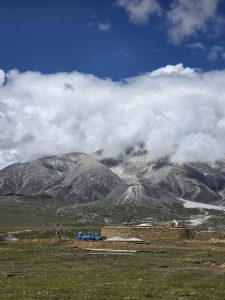 A high-mountain field featuring a stone structure and blue storage drums, backed by dark, barren mountain slopes partially covered by massive, low-hanging white clouds.