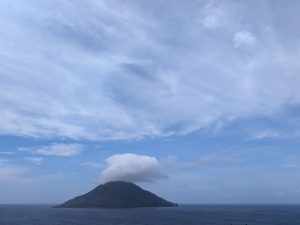 Cone-shaped Hachijo-kojima Island rising from a dark blue sea, with its summit covered by a round white cloud under a bright blue sky.