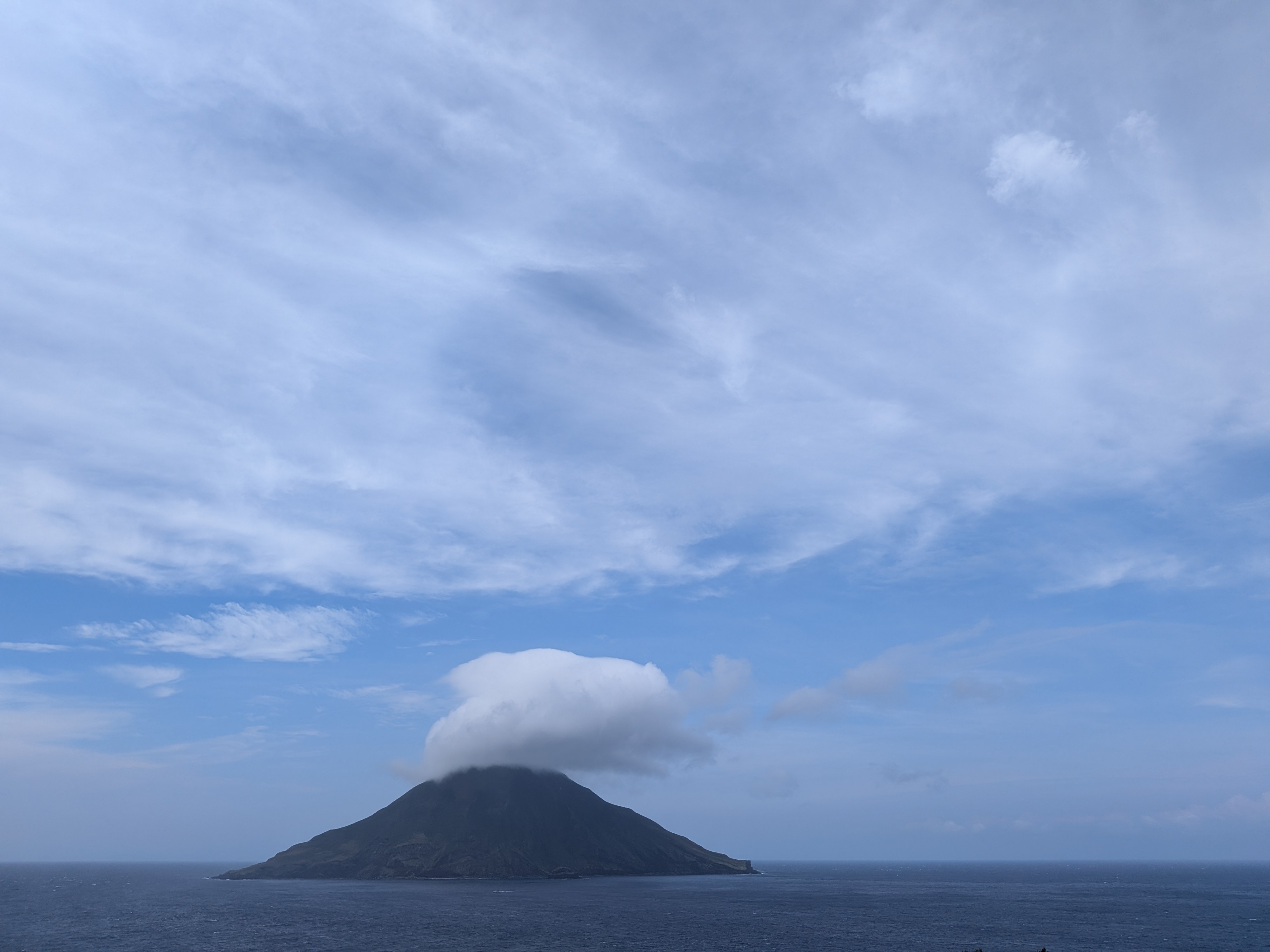 Cone-shaped Hachijo-kojima Island rising from a dark blue sea, with its summit covered by a round white cloud under a bright blue sky.