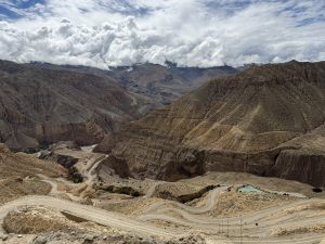 A high-angle perspective looking down at a winding mountain dirt road cutting through steep, arid gorge terrain toward a small turquoise pond.