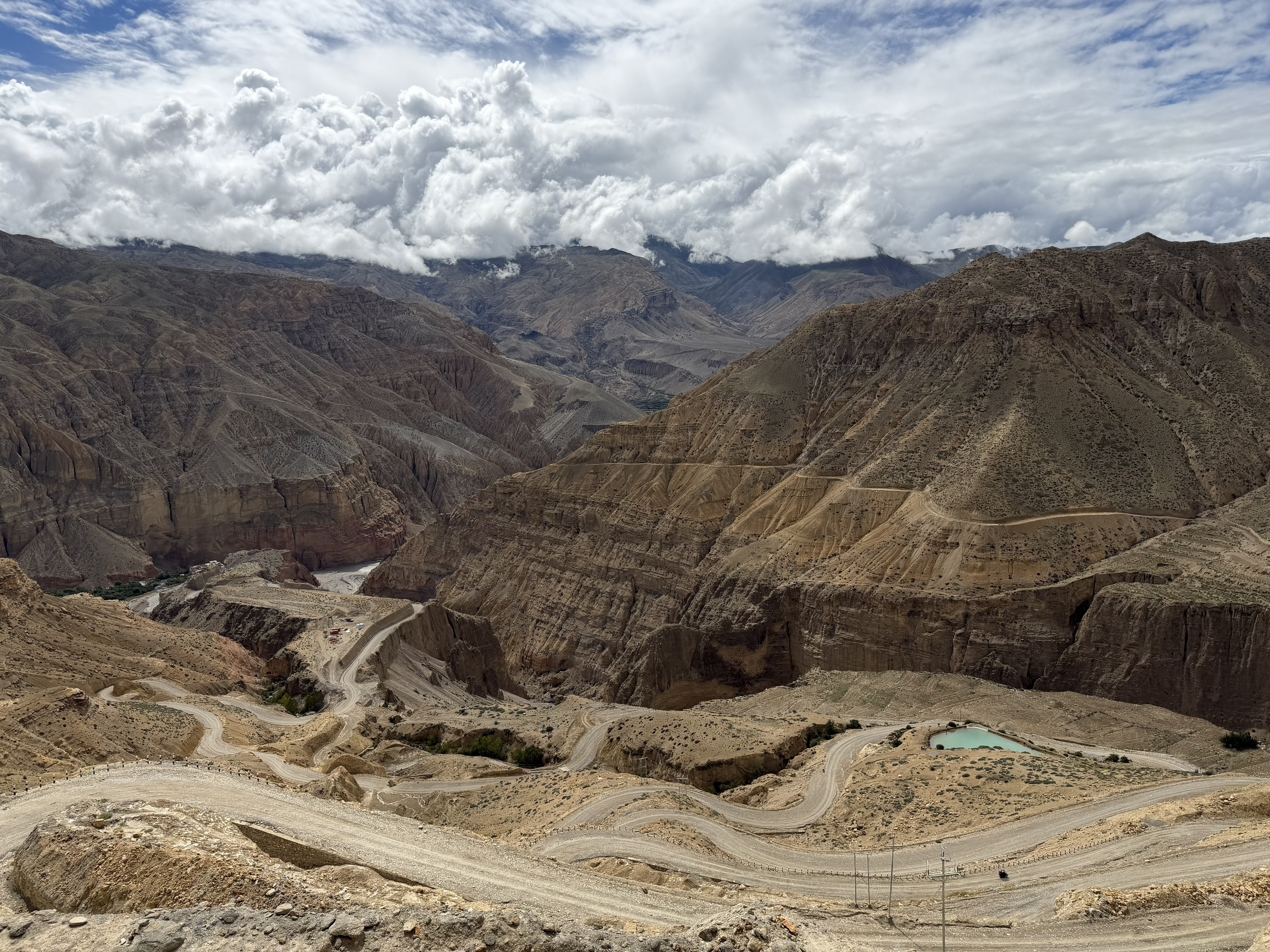 A high-angle perspective looking down at a winding mountain dirt road cutting through steep, arid gorge terrain toward a small turquoise pond.