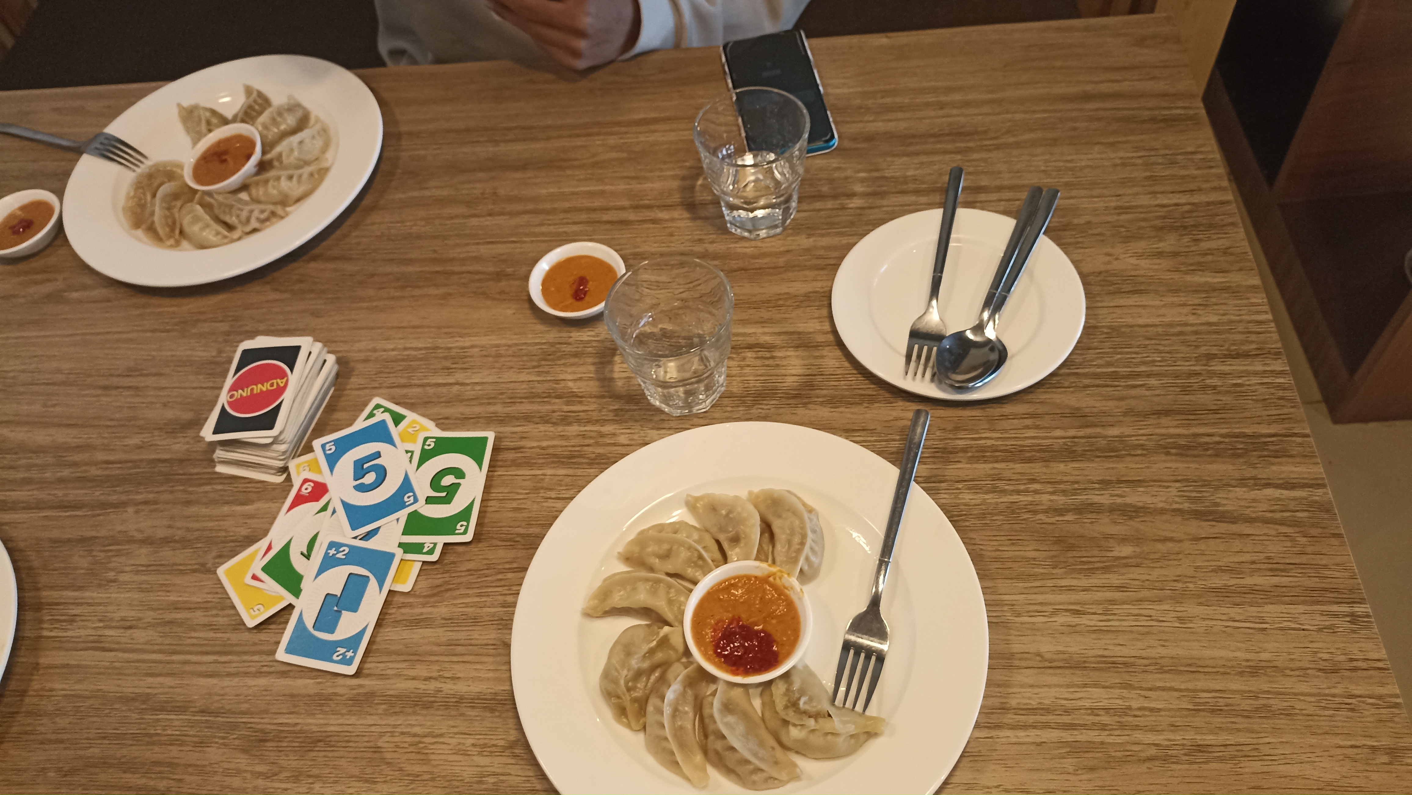 Plates of dumplings with dipping sauce, water glasses, utensils, and colorful playing cards on a wooden table.