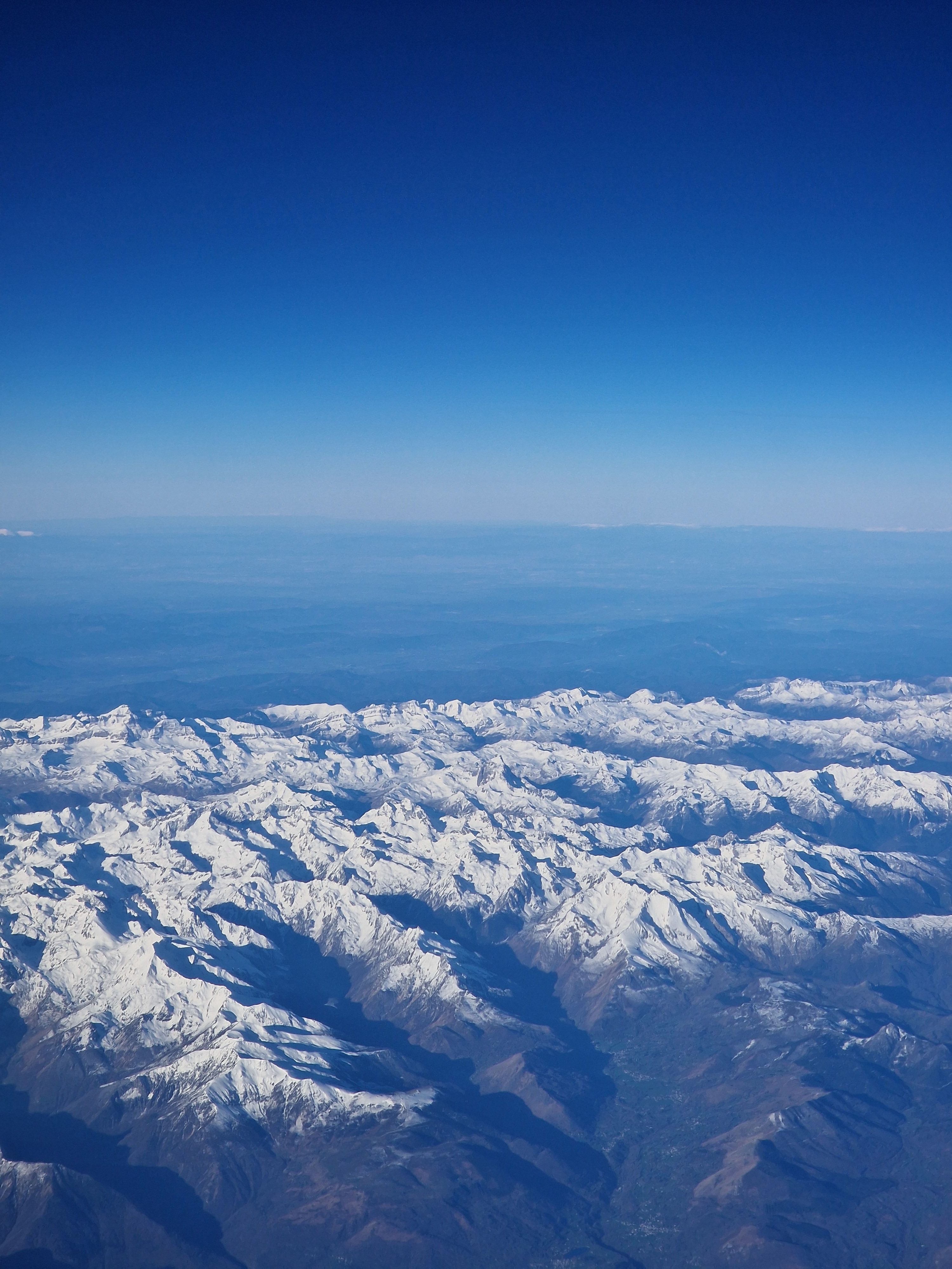 High-altitude aerial photograph looking down at a vast snow-covered mountain range stretching to the horizon beneath a deep blue gradient sky.