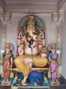 Lord Ganesha statue seated on a golden throne inside a temple, with smaller deities and a mouse at his feet.