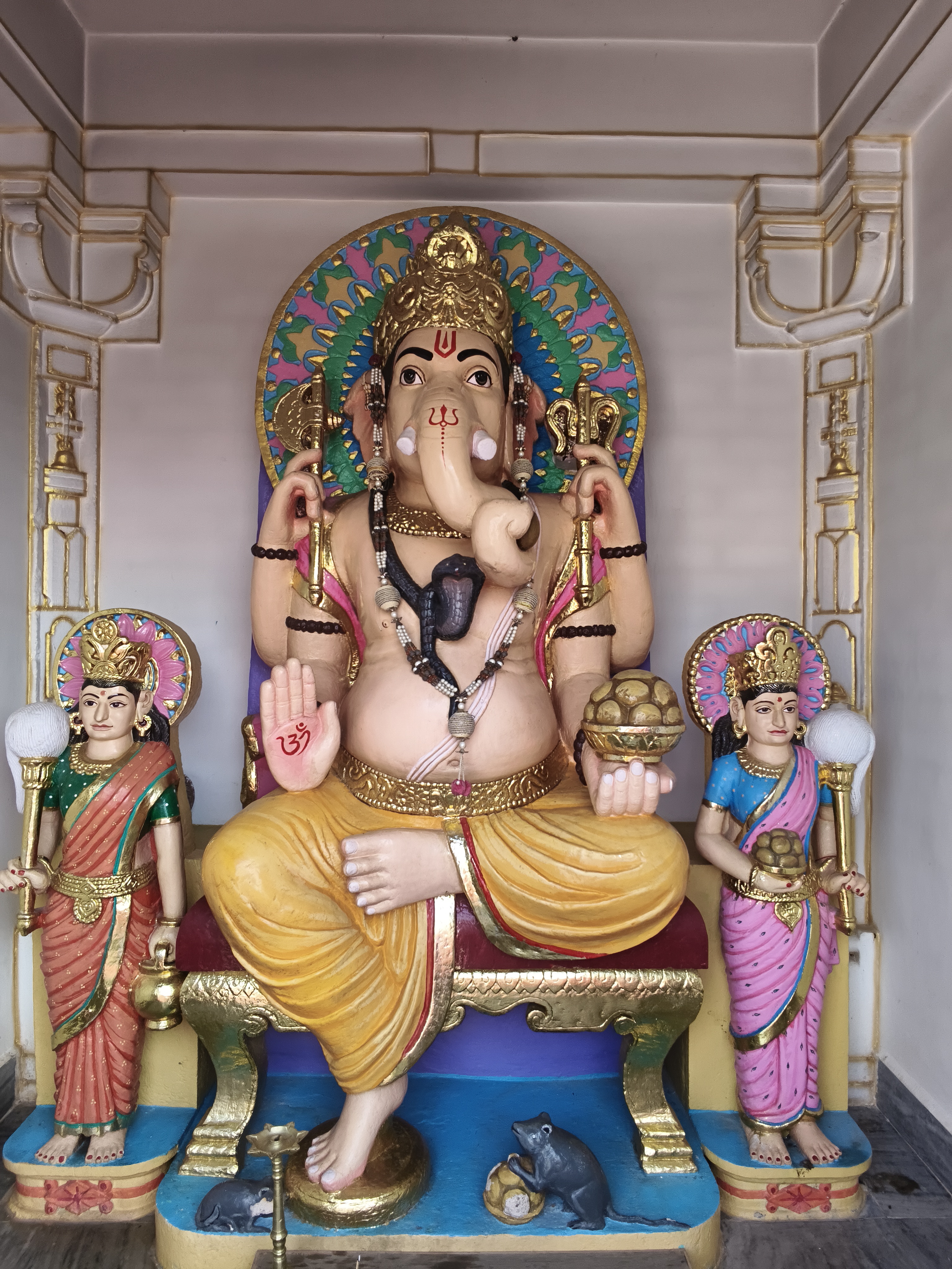 Lord Ganesha statue seated on a golden throne inside a temple, with smaller deities and a mouse at his feet.