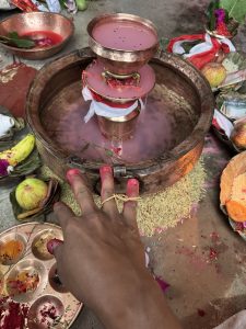 Hand with red-dyed fingertips and a grass ring reaching toward a copper ritual vessel filled with flowers and pinkish liquid, surrounded by fruits, grain, and rice offerings.