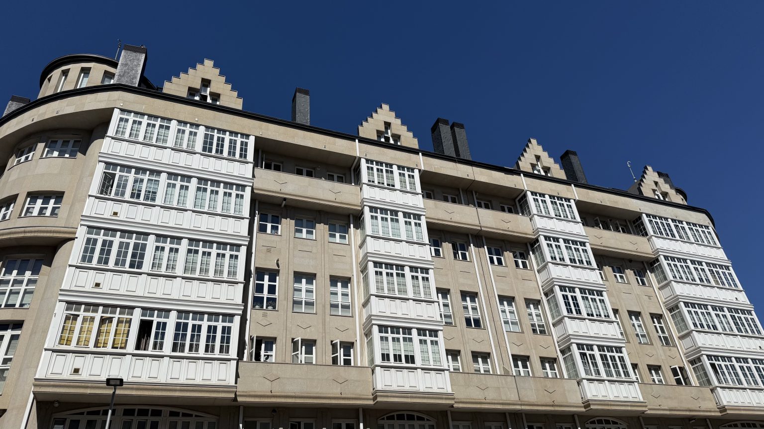 A long pale stone apartment building with columns of white glazed balconies and stepped gables, lit by hard sun under a deep blue sky.