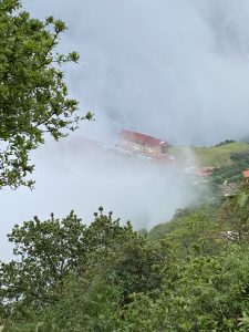 Misty hillside in Chandragiri with a red-roofed building surrounded by lush greenery and thick clouds.

