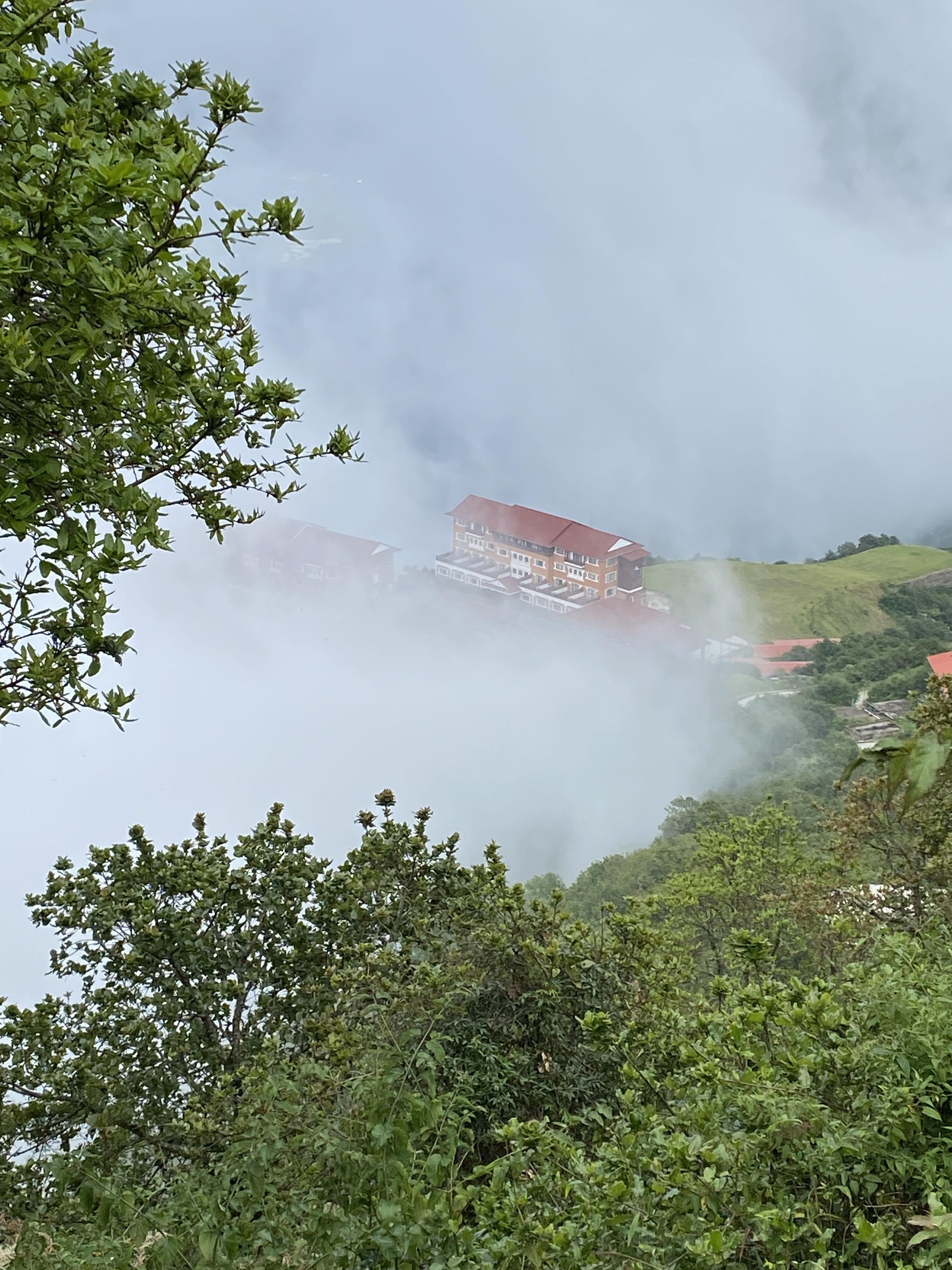 Misty hillside in Chandragiri with a red-roofed building surrounded by lush greenery and thick clouds.