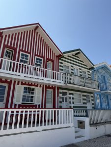 Three colorful houses in Costa Nova, Portugal, with red, green, and blue vertical stripes, white balconies, and dark brown shutters under a clear blue sky.