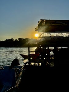 A riverboat silhouetted against the setting sun creates a striking scene, with golden light reflecting off the water. The low sun and calm ripples suggest a peaceful evening along the riverbank. 
