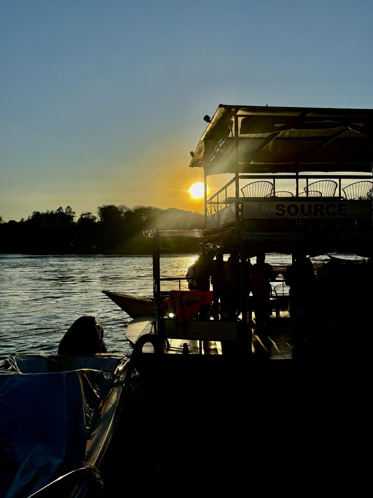 A riverboat silhouetted against the setting sun creates a striking scene, with golden light reflecting off the water. The low sun and calm ripples suggest a peaceful evening along the riverbank.