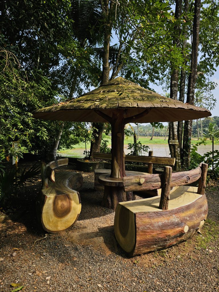 A rustic concrete seating shelter designed to resemble a wooden structure, featuring log-style benches and a broad mushroom-shaped roof, surrounded by tropical greenery and rice fields. Location: Ayamkudy, Kottayam, Kerala.
