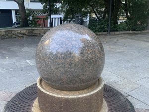 Polished granite sphere sculpture on a cylindrical pedestal in an urban plaza.