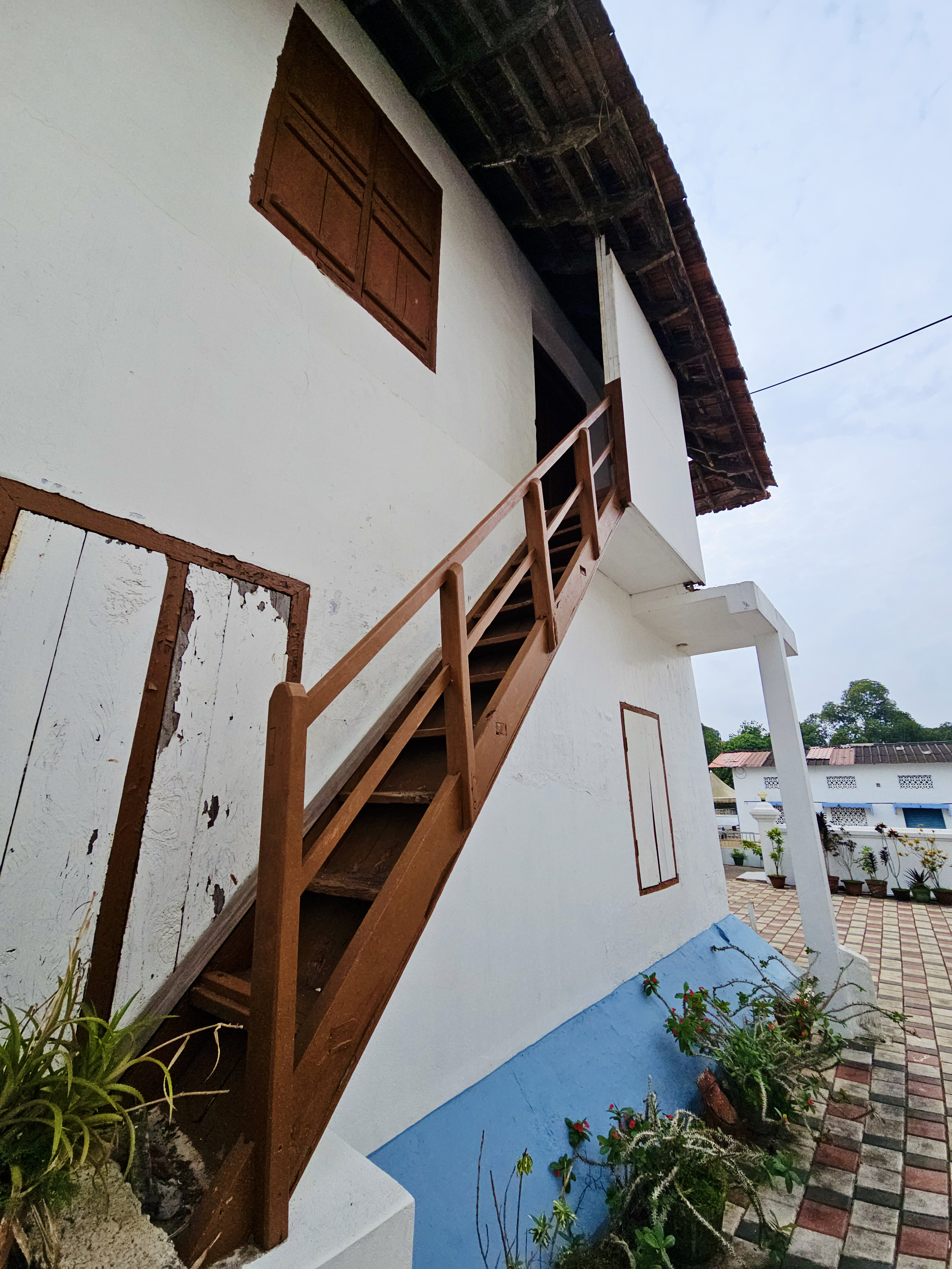 A traditional white building with a wooden exterior staircase, a tiled roof, and a blue-painted lower wall. The diagonal staircase creates an interesting architectural composition. Location: Ramapuram, Kottayam, Kerala.