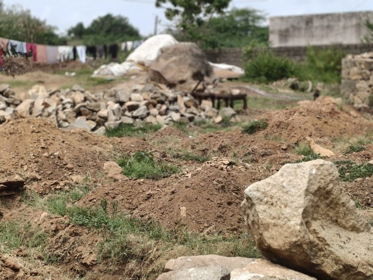 Rugged area with piles of dirt and rocks, colorful clothes hanging on a clothesline, and stone and concrete structures in the background.