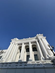 A low-angle shot looking up at the grand façade of a white neoclassical building featuring Corinthian-style columns and ornamental lampposts.