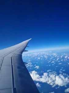 Aerial view from an airplane window looking along a silver wing over a sea of white scattered clouds and a deep blue sky.