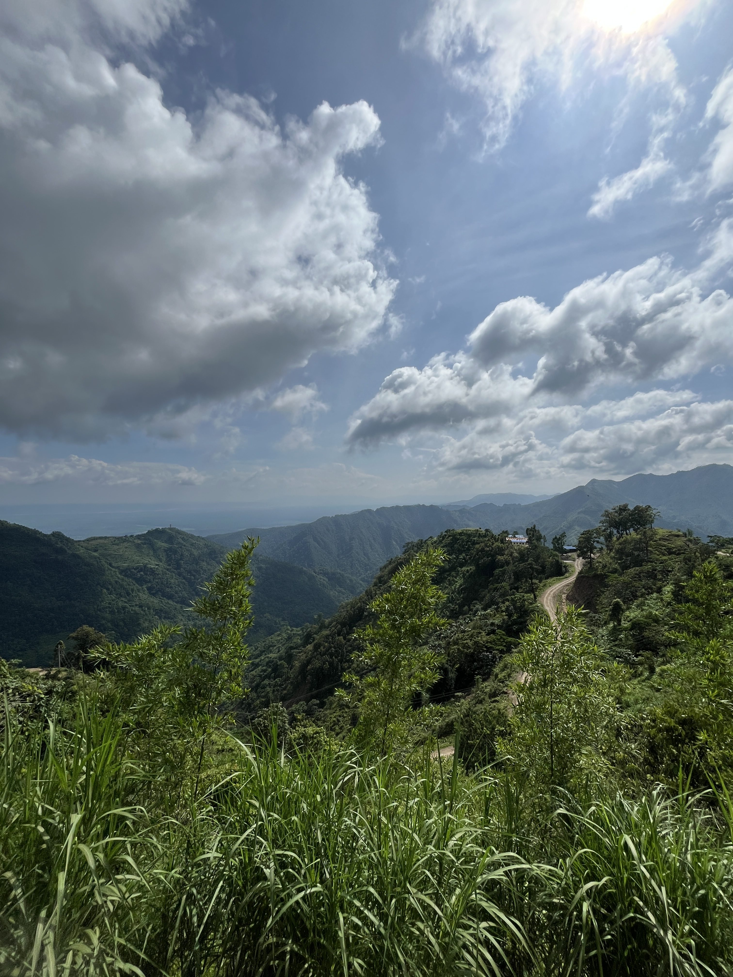 Lush green mountain ridges with a winding dirt road leading to a hilltop settlement under a cloudy blue sky.