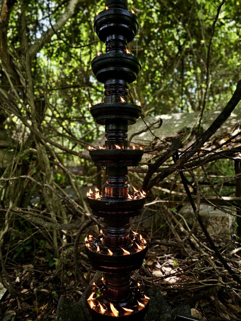 A tall traditional black oil lamp stands among the trees, with small flames glowing around each tier. The contrast between the dark lamp and the green forest creates a striking, atmospheric photograph. Location: Ayamkudy, Kottayam, Kerala.