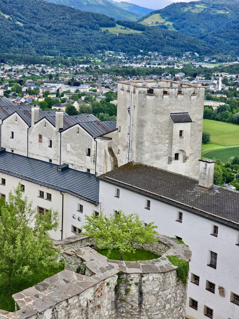 A historic white fortress complex overlooks Salzburg, with its weathered stone walls in the foreground and a picturesque town and forested Alpine hills stretching into the distance. Location: Salzburg, Austria.