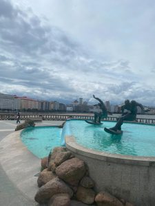 Large curved fountain with flowing water and bronze figures, surrounded by rocks, with city buildings in the background.
