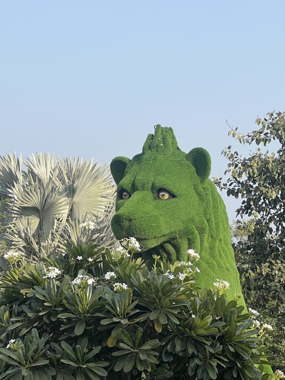 A lion-shaped green hedge sculpture (topiary) surrounded by palm leaves and white flowering plants, set against a clear blue sky.