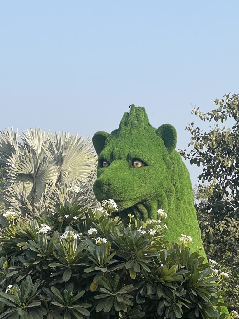 A lion-shaped green hedge sculpture (topiary) surrounded by palm leaves and white flowering plants, set against a clear blue sky.